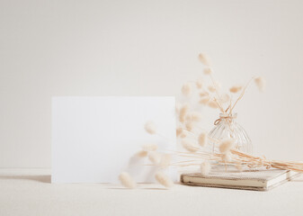 Blank white greeting card mock up.decoration with dried Lagurus ovatus flowers composition in modern glass vase and craft book  on  beige table and cement wall background
