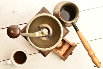 One wooden manual coffee grinder with a cup of coffee and cezve, close-up, on a white wooden table.