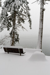 A single snow covered bench looking out over the water in a snow storm.