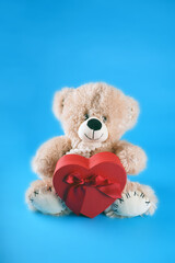 Close up of cute teddy bear with red gift box in form of heart. Soft plush toy with present on blue background. Concept of holidays, presents and good mood.