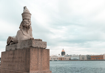 Saint-Petersburg, Russia, 22 August 2020: Sphinx at the Universitetskaya Embankment of Neva river on background of old architectural buildings and Saint Isaac's Cathedral in cloudy weather.