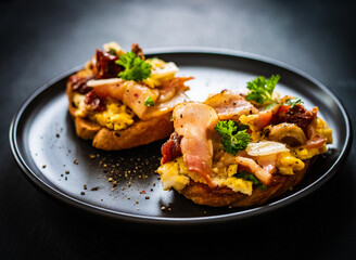 Delicious breakfast - scrambled eggs with bacon and champignons and sun-dried tomatoes on toasted bread on black table
