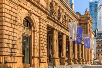 historic building of the stock exchange in Frankfurt with flags above the entrance. brown colored commercial building in baroque style. Skyscrapers in the background. Trees in the foreground in spring - obrazy, fototapety, plakaty