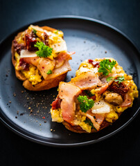 Delicious breakfast - scrambled eggs with bacon and champignons and sun-dried tomatoes on toasted bread on black table
