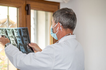 Doctor wearing face mask looking at x-ray backlit at the room window