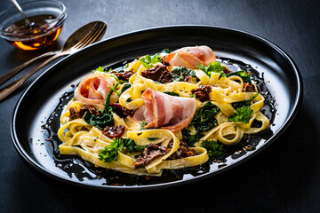 Tagliatelle with bacon, sun-dried tomatoes and spinach in alfredo sauce on black wooden table 
