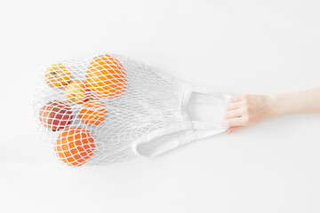 Fruits in white mesh bag with hand flat lay on white background top view. Eco friendly, reusable shopping bag. Oranges, apples, garnet in cotton knitted string bag. Zero waste concept.