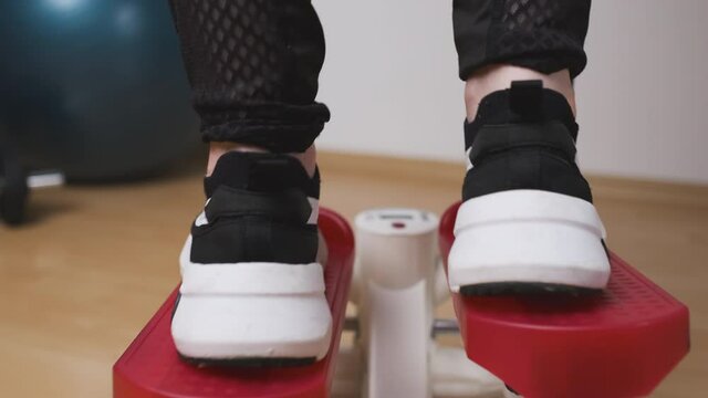 Back view of female legs doing exercises on the twist stepper in sneakers and sportswear at home during lockdown. Workout at home 