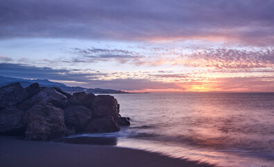 Picturesque scene of the purple toned sunset above sea waters seen through the rocky shore