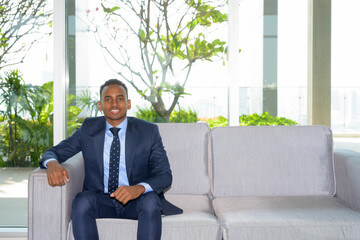 Confident happy African businessman wearing suit and sitting on sofa at modern office