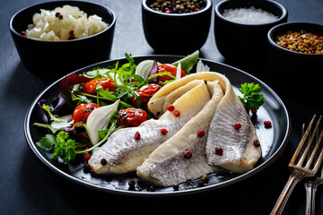 Marinated herrings with roast cherry tomatoes and fresh vegetable leaves on black table

