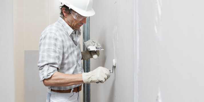 man drywall worker or plasterer putting plaster on plasterboard wall using a trowel and a spatula, fill the screw holes, wearing white hardhat, work gloves and safety glasses.