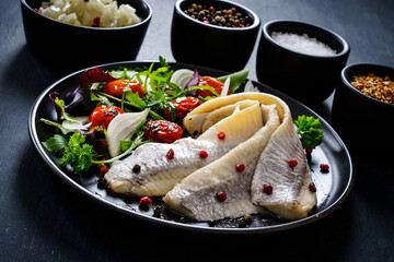 Marinated herrings with roast cherry tomatoes and fresh vegetable leaves on black table

