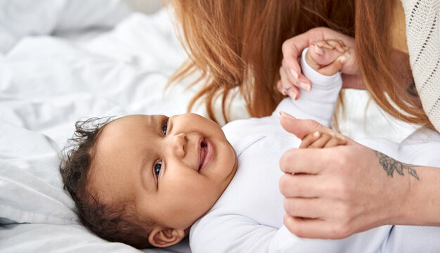 Happy Cute Little Funny African American Baby Daughter Playing With Caucasian Mom In Bed. Diverse Mother And Infant Child Daughter Having Fun Together Enjoying Soft Comfortable Textile At Home.