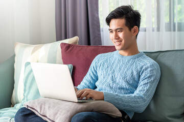 Happy of a leisurely day of a young Asian man watching his laptop, sitting on the sofa at home.