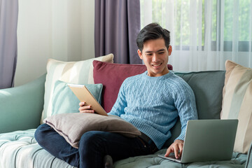 Young Asian man on a leisurely day sitting home on sofa using laptop and tablet on the couch.