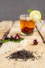 black tea in a glass cup with mint cherries and lemon on a wooden table next to fresh cherries and sprinkled tea leaves.