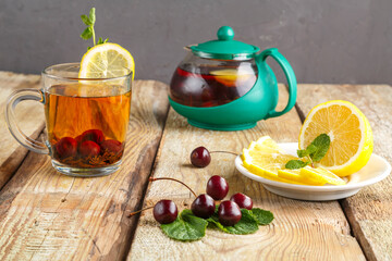black tea in a glass cup with mint cherries and lemon on a wooden table next to fresh cherries and a teapot.