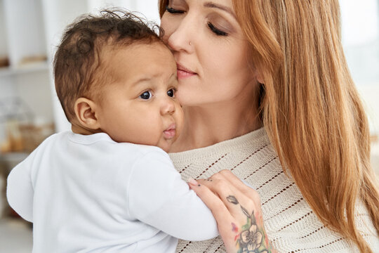 Loving young caucasian mother hugging cute infant african american baby daughter expressing single parent love, tenderness and care. Diverse mum and adorable ethnic little child girl bonding.