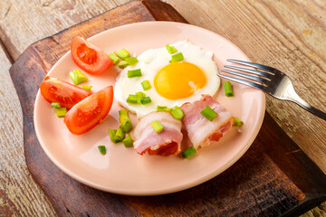 Fried eggs with bacon decorated with tomato and green onion on a wooden board.