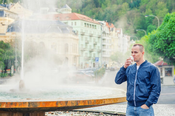 Young man tourist with blue jacket look at camera and drink therapeutic mineral water from cup,...