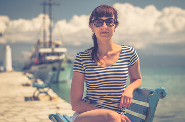 Young beautiful girl with striped t-shirt and sunglasses smile and sit on bench in pier, white ship on Toroneos gulf blue turquoise water in Halkidiki in sunny summer day background, close-up view