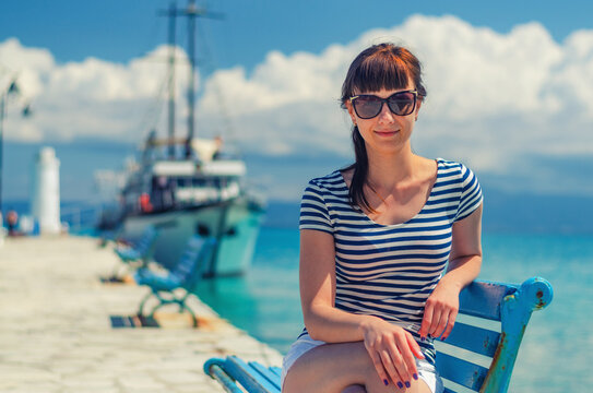 Close-up view of young beautiful girl with striped shirt and sunglasses smile and sit on bench in pier, white ship on Toroneos gulf turquoise water in Halkidiki in sunny day, vacation in Greece