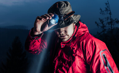 Fototapeta Photo of a male hiker in red jacket and panama putting on head mount flashlight on dark mountain forest background. obraz