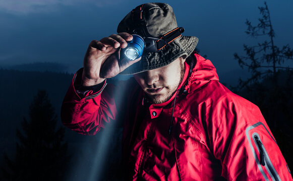Photo of a male hiker in red jacket and panama putting on head mount flashlight on dark mountain forest background.