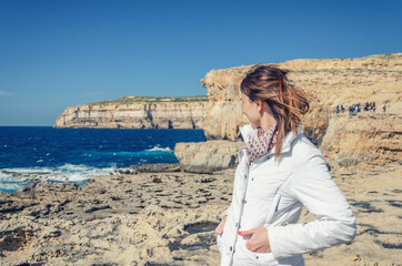 Fototapeta na wymiar Young woman traveler with white jacket keep hands in pocket, looking away distance on stone rocks of Dwejra Bay, collapsed Azure Window in Gozo island of Mediterranean sea background, Malta vacation