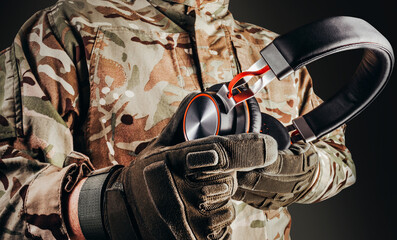 Photo of soldier in camouflaged uniform and tactical gloves holding wireless headphones on black...