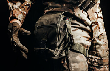 Photo of soldier in camouflaged uniform and tactical gloves using leg bag on black background.