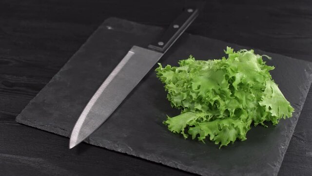 green fresh lettuce and wahed steel chefs knife shiny reflections rotating on black stone plate close up view