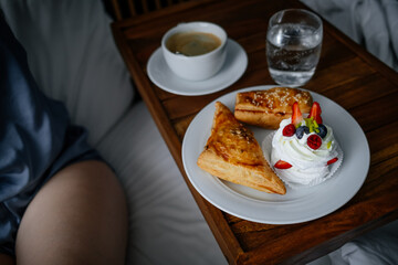 Female enjoying a cup of coffee with delicious pastries on a board on the bed