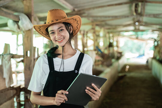 close up of a smiling woman wearing a cowboy hat carrying a digital tablet in the background of a cow farm stable