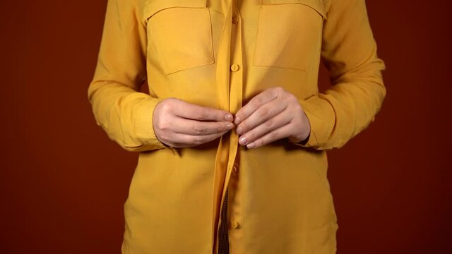 Front view on body part of young female in yellow unbuttoned blouse. Close up of unrecognizable woman buttons her blouse on orange background.