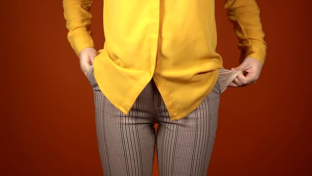 Close up of unrecognizable woman put her hands in her pockets of pants. Front view on body part of young female showing her empty pockets on orange background.