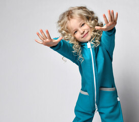 smiling happy girl in warm trendy jumpsuit, trendy comfortable sportswear, reaching for portrait in studio. Pretty child beauty model with blond curly hair. Happy childhood