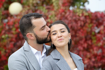 Stylish couple in gray coats gently hugging on the background of autumn leaves. Happy sensual wedding couple embracing. Romantic moments of newlyweds.