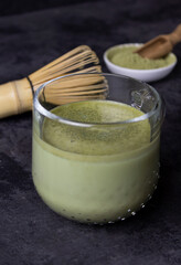 Glass cup with japanese matcha tea on a black surface, bamboo whisk for whipping and green matcha powder on the background