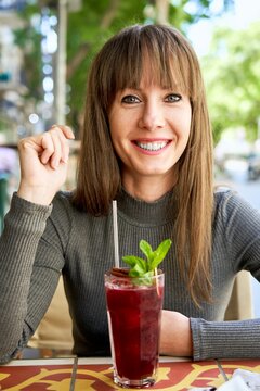Portrait of attractive happy young woman looking at camera on cafeteria terrace drinking cocktail.