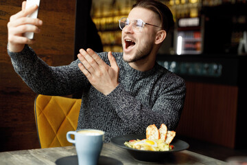 Beautiful smiling male in cafe drink coffee and eating salad. Man in casual clothes and glasses with smartphone in hands. Blogger influencer looking at camera make video conference call recordin.