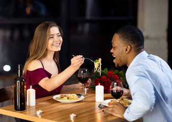 Cheerful interracial couple having fun at dinner in restaurant, woman feeding boyfriend