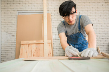 Young man carpenter wearing safety goggle using pencil and Iron ruler draw line on plank wood for...