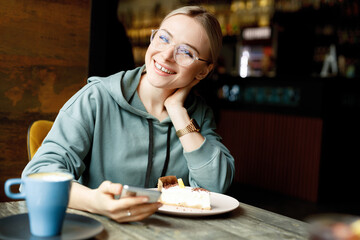 Beautiful smiling female in cafe drink coffee and eating cake. Young woman in casual clothes and glasses with smartphone in hands.