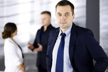 Portrait of a self-confident middle aged businessman in a blue suit, standing in a modern office with his colleagues at the background. Concept of business success