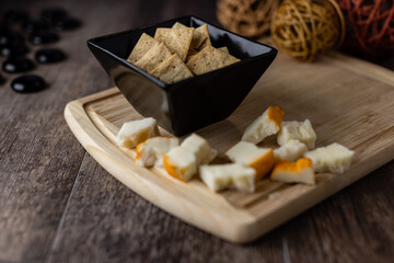 A black bowl of snack crackers and assorted cheese on a cutting board sitting on a wooden table top