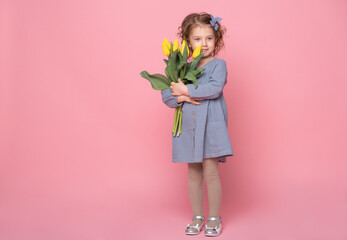 beautiful little blonde girl in blu dress with bouquet of yellow tulips on pink background with space for text