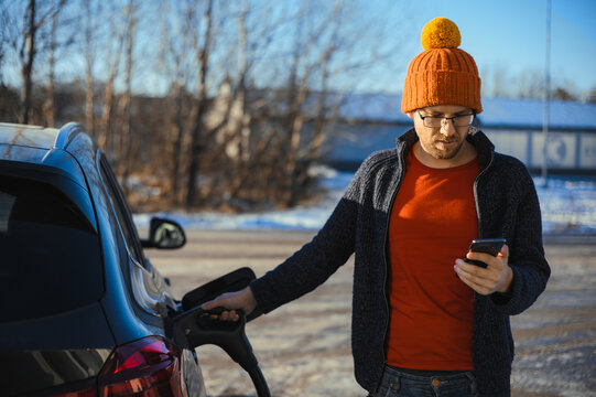 Nerdy Man In The Orange Hat With Phone Is Charging The Electric Car On The Charge Station.