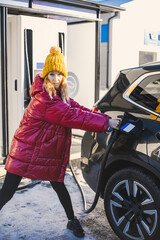 Fototapeta na wymiar Beautiful girl in the red jacket and yellow hat with phone is charging the electric car on the charge station.